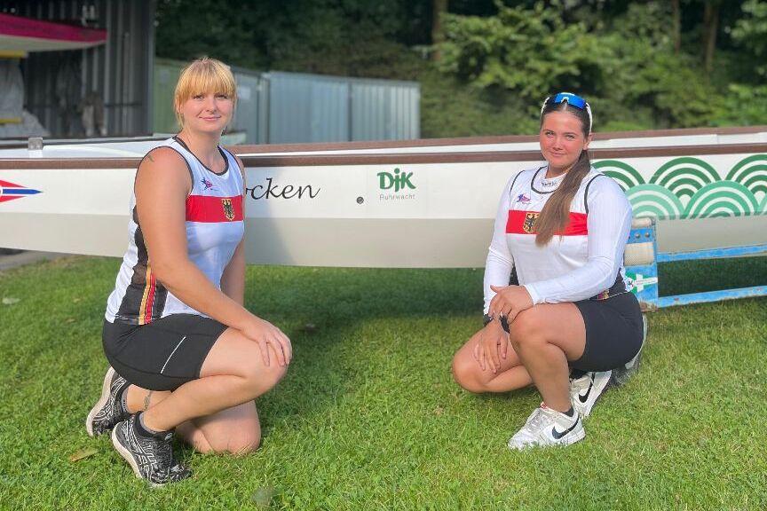 Zwei Ruderinnen knien auf gr&uuml;nem Gras neben einem Ruderboot, umgeben von bunten Fahnen und B&auml;umen.