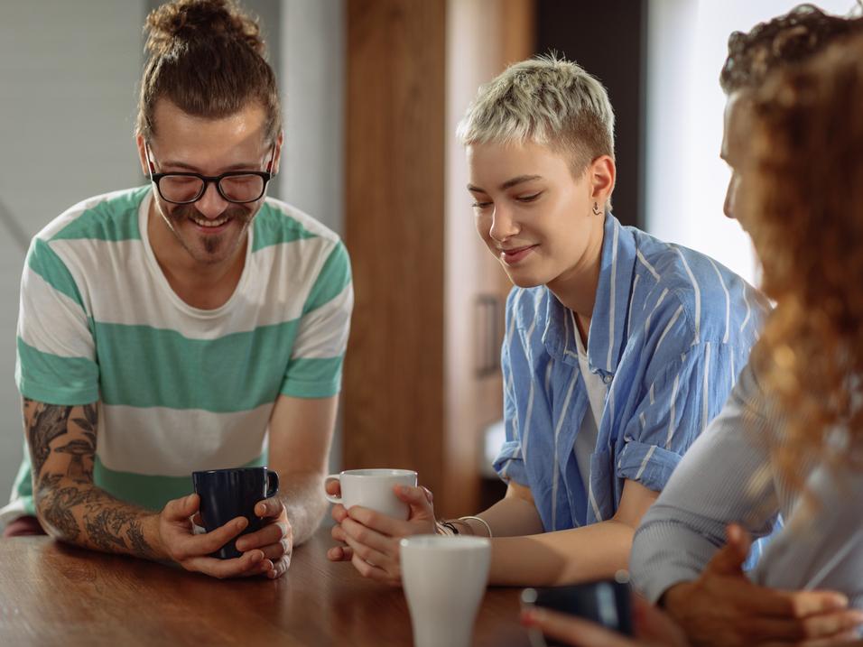 Vier Personen sitzen an einem Tisch und betrachten l&auml;chelnd ein Smartphone, w&auml;hrend sie Tassen in den H&auml;nden halten.