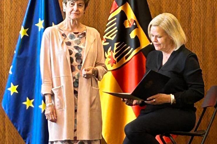 Zwei Frauen stehen auf einer B&uuml;hne, umgeben von Europaflagge und Bundesflagge, eine h&auml;lt eine Mappe.