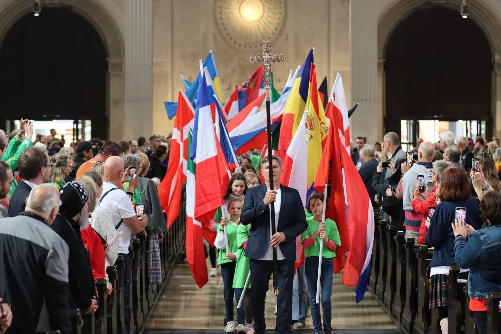 Fahnentr&auml;ger und Kinder mit Flaggen marschieren in einer festlichen Halle, umgeben von jubelnden Zuschauern.