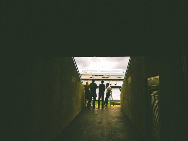 Silhouette von vier Personen im Stadion-Tunnel, Blick auf das Spielfeld und den bew&ouml;lkten Himmel.