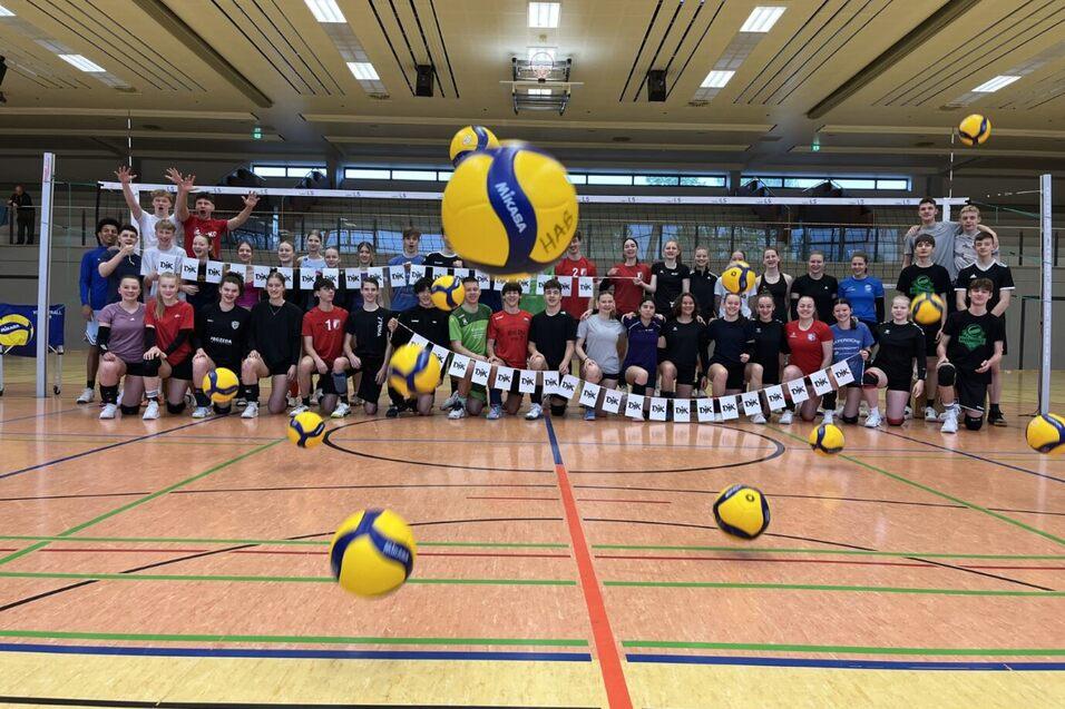 Gruppenfoto von Volleyballspielern in einer Sporthalle, mit mehreren B&auml;llen in der Luft und bunten Trikots.