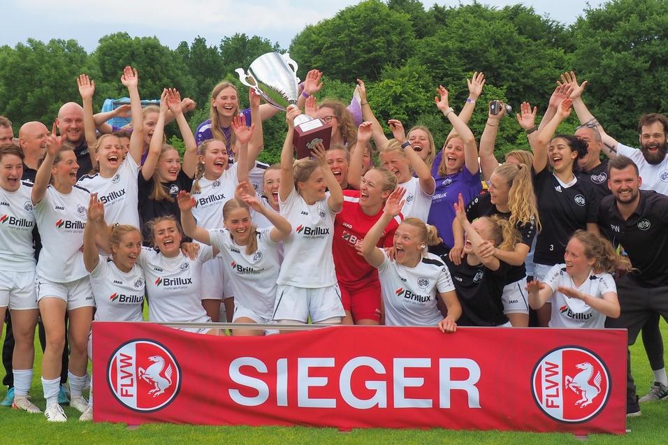 Mannschaft feiert mit Pokal und Banner "SIEGER" auf dem Fu&szlig;ballplatz, umgeben von jubelnden Spielerinnen und Unterst&uuml;tzern.