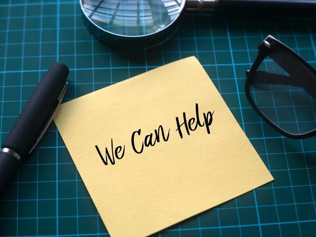 Gelber Notizzettel mit der Aufschrift „We Can Help“, daneben ein Kugelschreiber, eine Lupe und eine Brille auf einem grünen Schneidebrett.