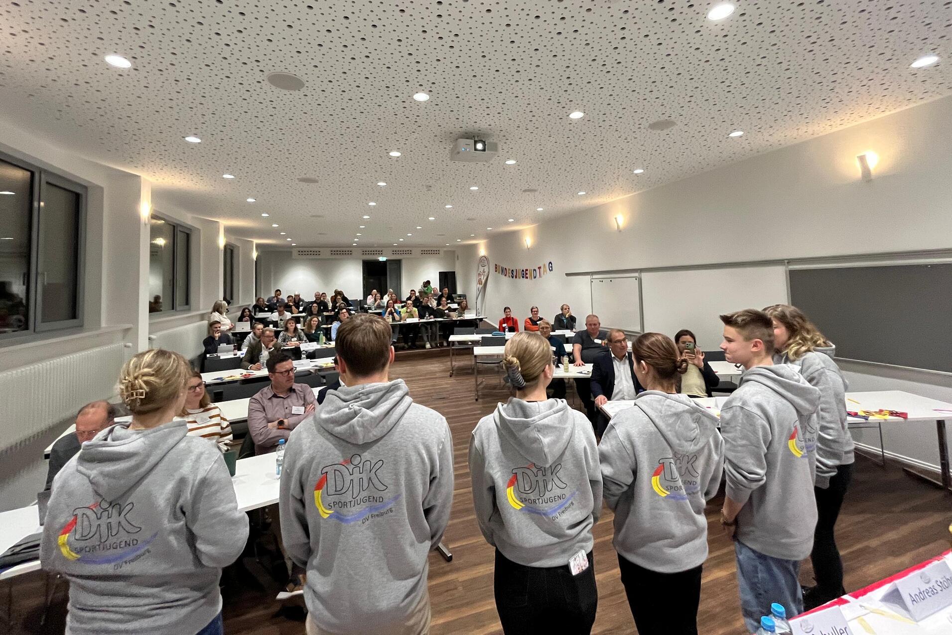 Sechs Personen in grauen Hoodies stehen vor einer Gruppe von Teilnehmern in einem modernen Seminarraum.