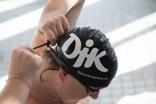 Sportler zieht eine schwarze Badekappe mit Aufschrift &bdquo;DJK Sportverband&ldquo; an, w&auml;hrend er sich auf dem Schwimmbadrand vorbereitet.