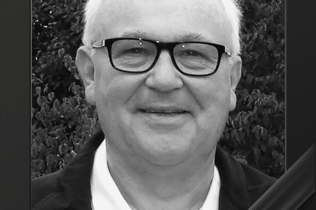 Schwarz-wei&szlig;e Portraitaufnahme eines &auml;lteren Mannes mit Brille, l&auml;chelnd vor einem gr&uuml;nen Hintergrund.