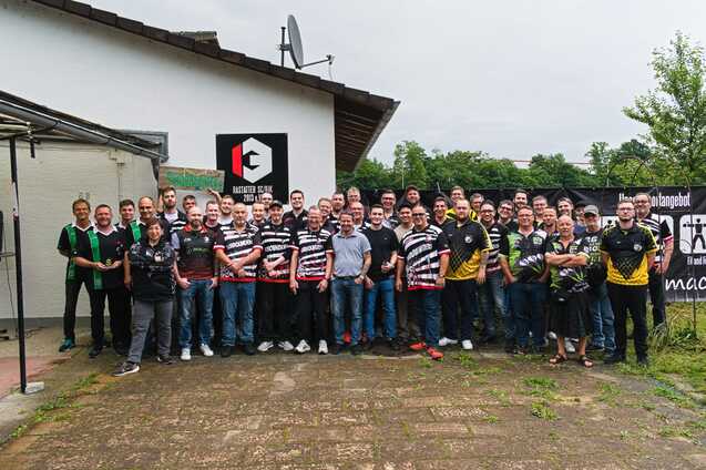 Gruppenfoto von 50 Personen in Team-Outfits vor einem Geb&auml;ude mit Banner und Logos im Freien.