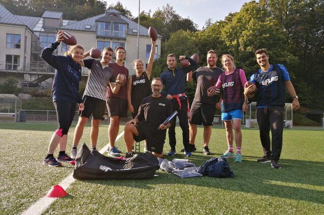 Eine Gruppe von neun Personen h&auml;lt American Footballs auf einem Sportplatz mit Kunstrasen. Im Hintergrund sind B&auml;ume und ein Geb&auml;ude.