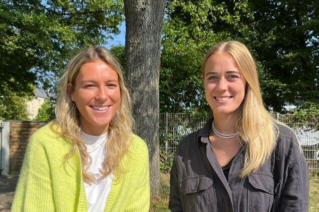 Zwei Frauen l&auml;cheln vor einem Baum und einer gr&uuml;nen Landschaft, beide tragen l&auml;ssige Kleidung.