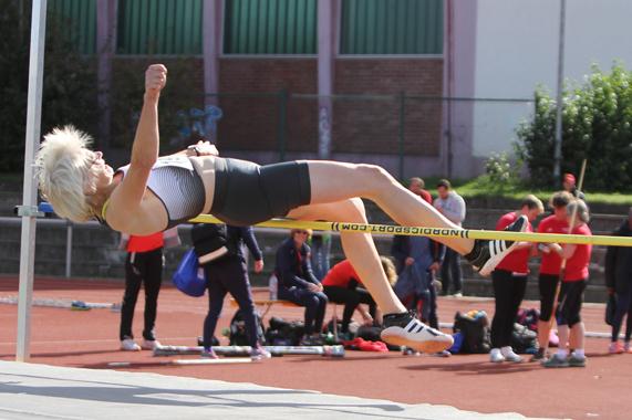 Sportlerin springt &uuml;ber eine Hochsprunglatte, w&auml;hrend Zuschauer im Hintergrund ihre Technik beobachten.