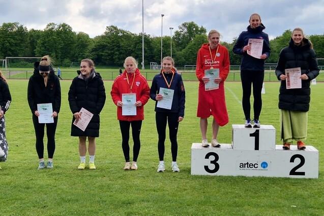 Sieben Frauen stehen auf einem Siegertreppchen mit Medaillen und Urkunden, auf einem Sportplatz umgeben von B&auml;umen.