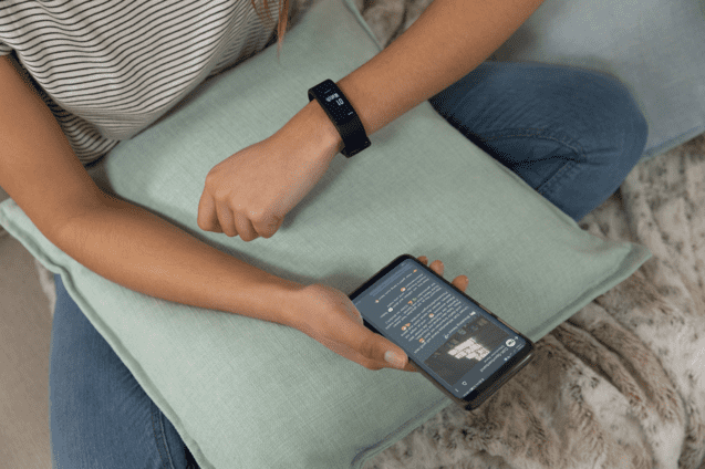 Junge Frau mit Fitness-Armband schaut auf ihr Smartphone, w&auml;hrend sie auf einem Kissen sitzt.