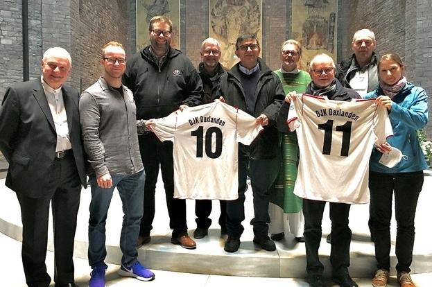 Gruppe von acht M&auml;nnern und einer Frau h&auml;lt zwei Fu&szlig;balltrikots mit den Zahlen 10 und 11 in einem Raum mit Mosaiken.