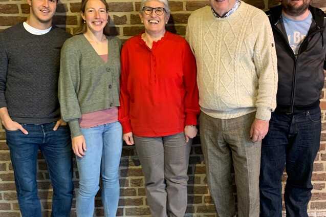 F&uuml;nf Personen stehen l&auml;chelnd vor einer Ziegelsteinwand, die Frauen in der Mitte tr&auml;gt ein rotes Oberteil.