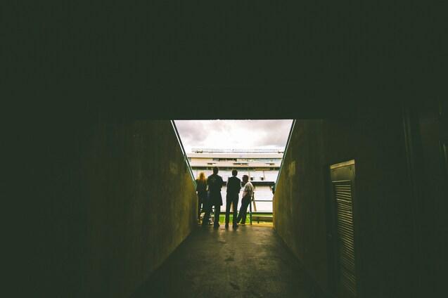 Silhouette von vier Personen im Stadion-Tunnel, Blick auf das Spielfeld und den bew&ouml;lkten Himmel.