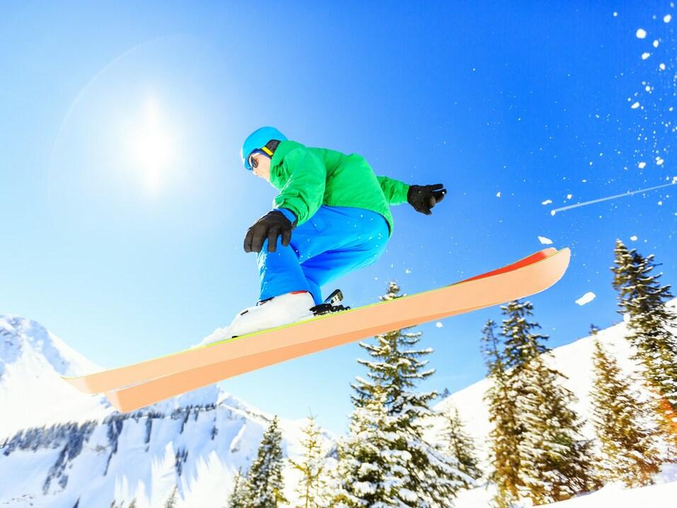 Ein Snowboarder springt über einen Schnee bedeckten Hang unter klarem blauen Himmel und strahlender Sonne.
