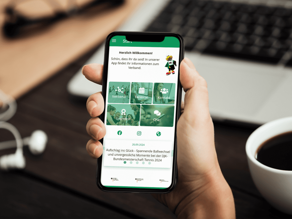 Hand h&auml;lt Smartphone mit App-Startseite, davor steht eine Tasse Kaffee und Kabelkopfh&ouml;rer auf einem Tisch.