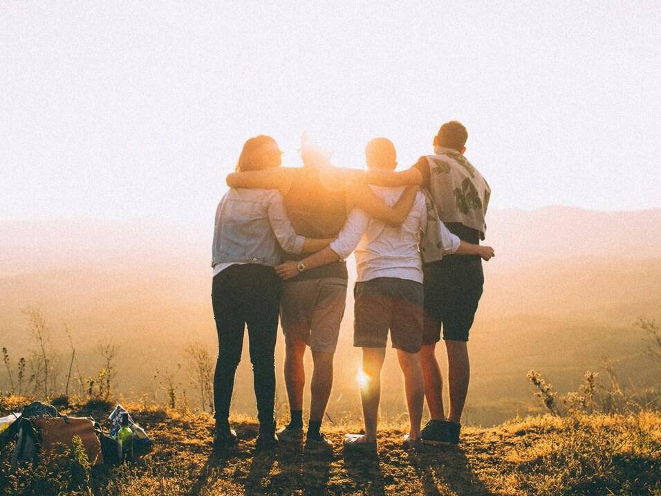 Gruppe von vier Menschen steht umarmt auf einem H&uuml;gel, w&auml;hrend die Sonne im Hintergrund untergeht.
