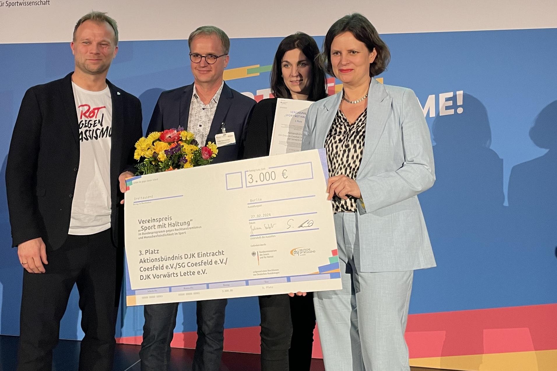 Vier Personen halten einen gro&szlig;en Scheck &uuml;ber 3.000 Euro, w&auml;hrend im Hintergrund ein Bildschirm den dritten Platz f&uuml;r &bdquo;Aktionsb&uuml;ndnis&ldquo; zeigt.