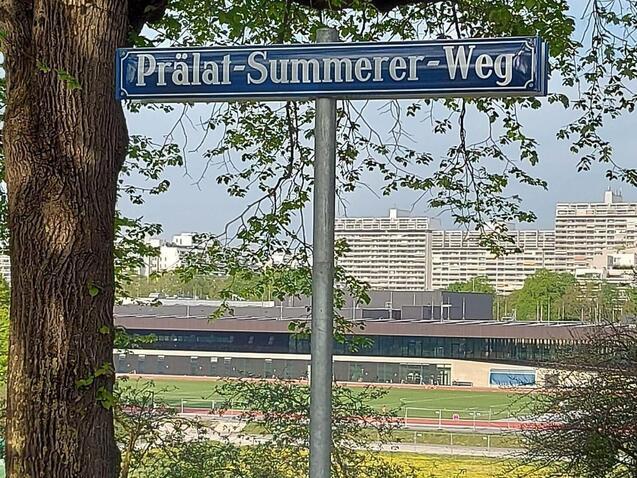 Stra&szlig;enschild &bdquo;Pr&auml;lat-Summerer-Weg&ldquo; vor B&auml;umen und einer Wiese, mit modernen Geb&auml;uden im Hintergrund.