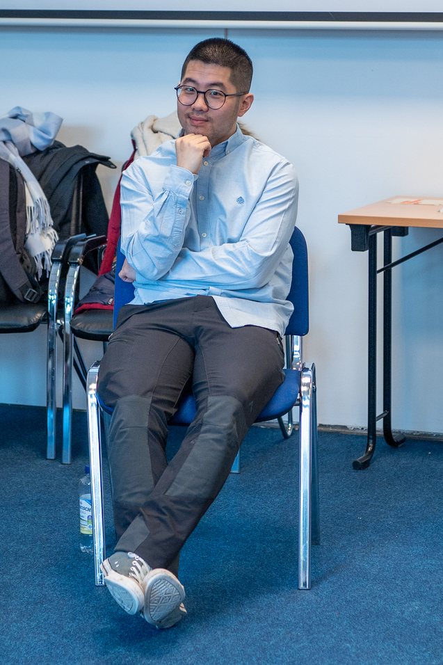 Junger Mann mit Brille in einer hellblauen Hemd sitzt entspannt auf einem Stuhl in einem Seminarraum.