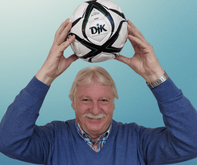 L&auml;chelnder Mann h&auml;lt einen Fu&szlig;ball &uuml;ber seinem Kopf, vor einem einfarbigen Hintergrund.