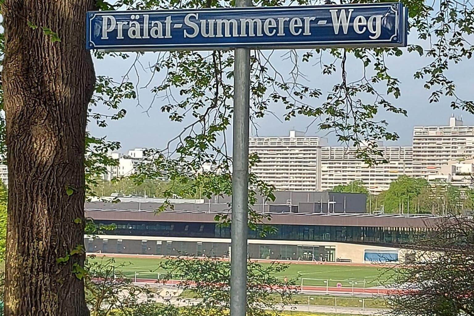 Stra&szlig;enschild &bdquo;Pr&auml;lat-Summerer-Weg&ldquo; vor B&auml;umen und einer Wiese, mit modernen Geb&auml;uden im Hintergrund.