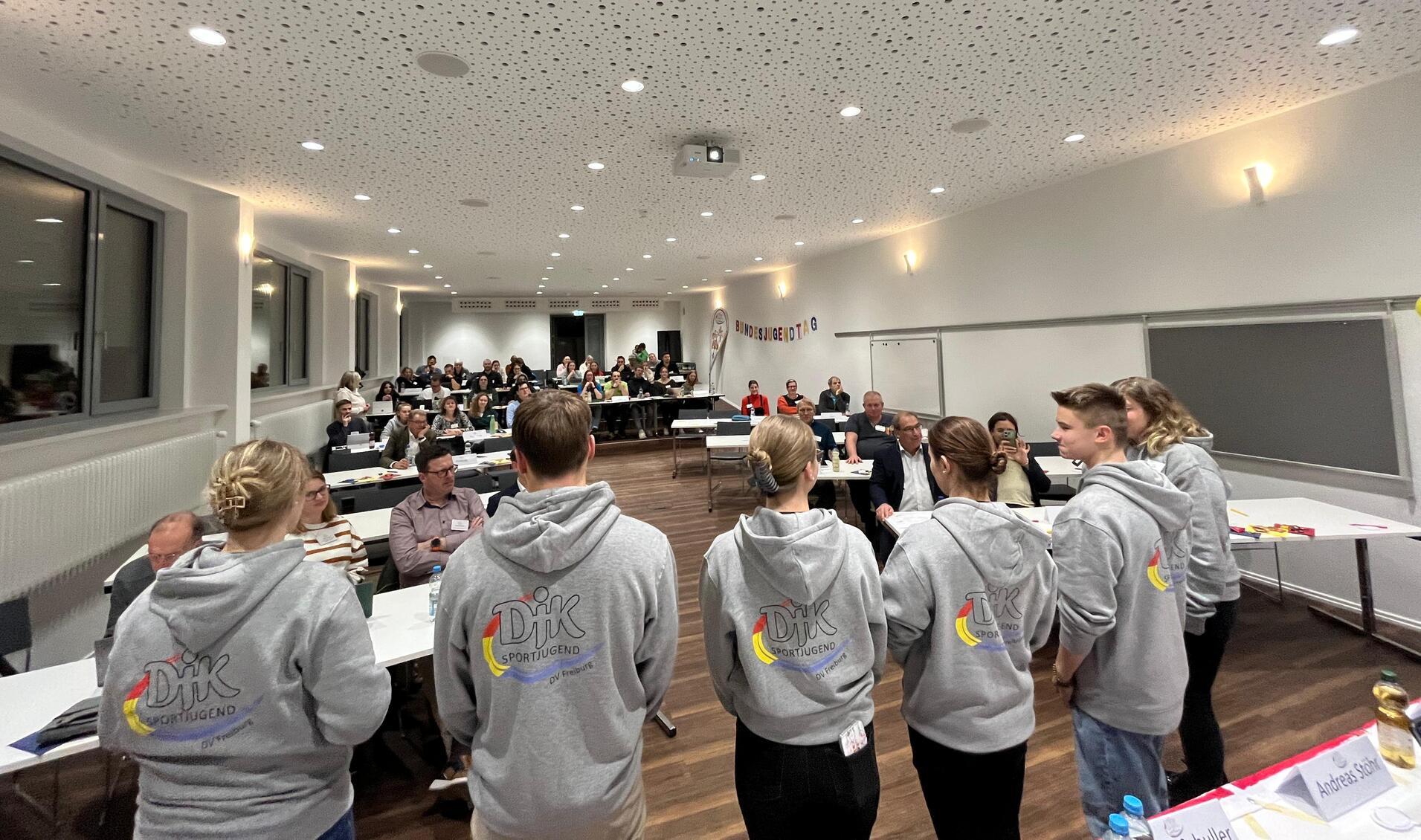 Sechs Personen in grauen Hoodies stehen vor einer Gruppe von Teilnehmern in einem modernen Seminarraum.