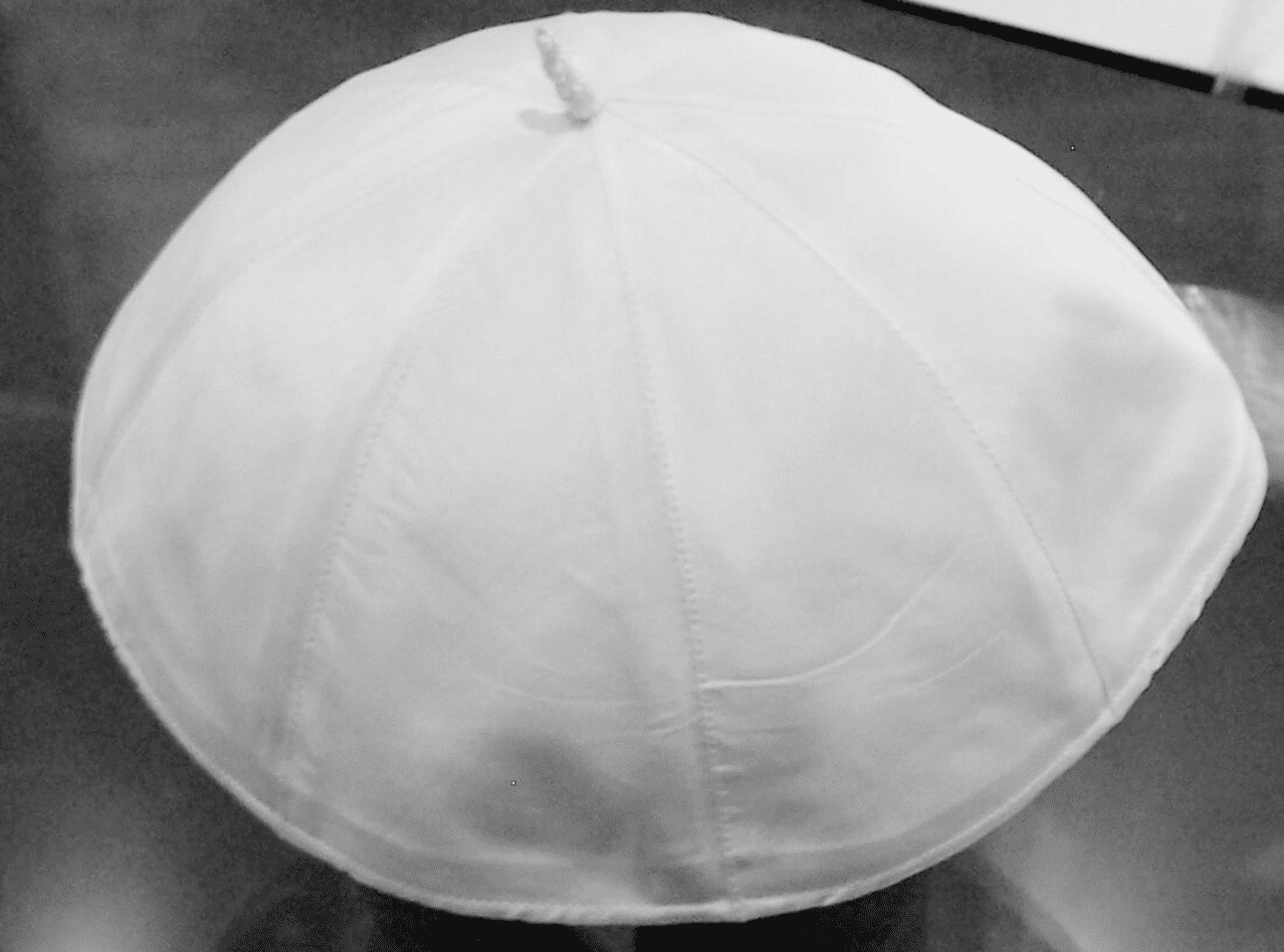 Wei&szlig;e Kappe aus Stoff, leicht gew&ouml;lbt mit einer kleinen Spitze oben, auf einem dunklen Untergrund.