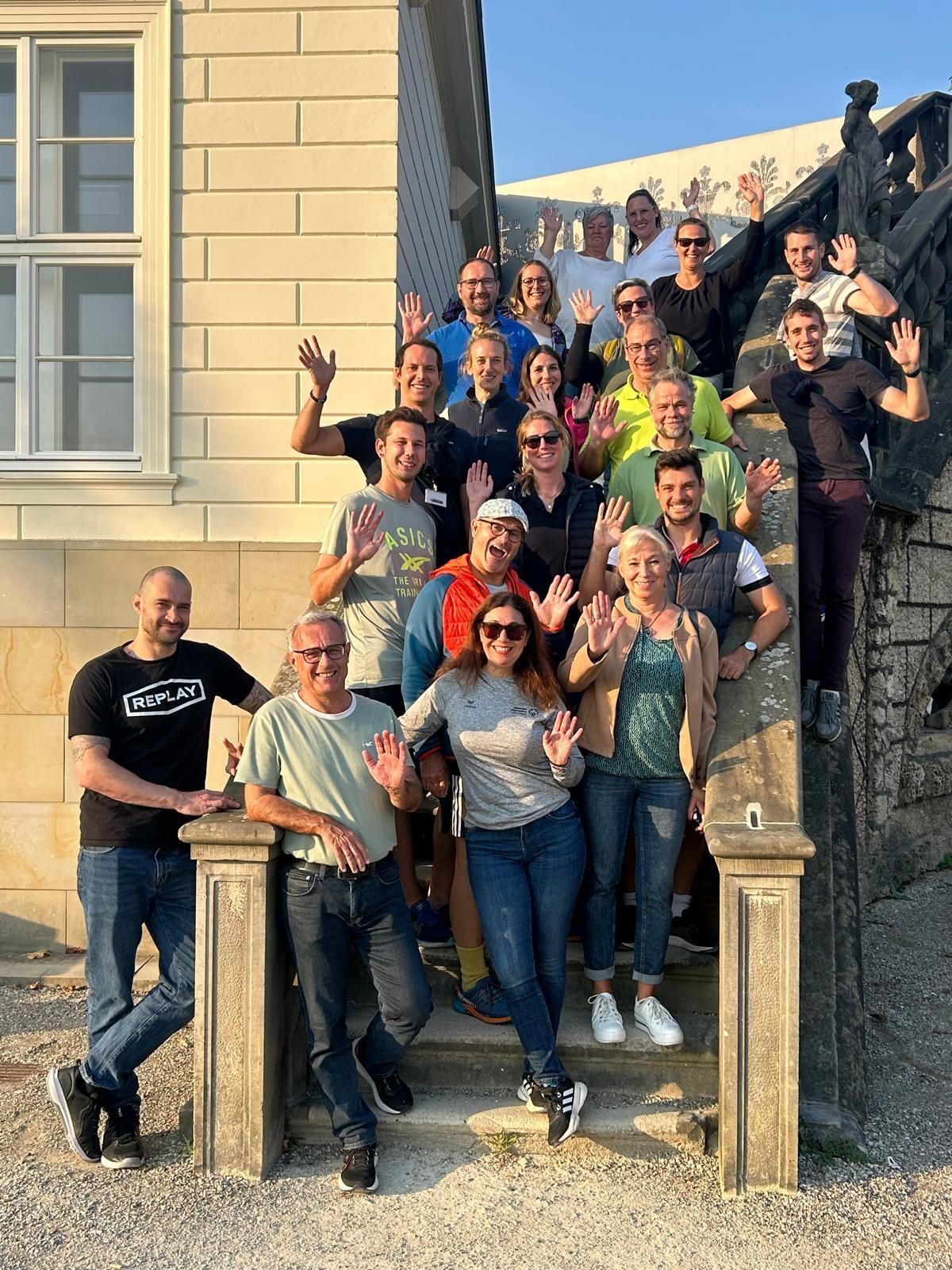 Gruppe von 24 Personen, die fr&ouml;hlich auf einer Treppe vor einem historischen Geb&auml;ude posiert und die H&auml;nde heben.