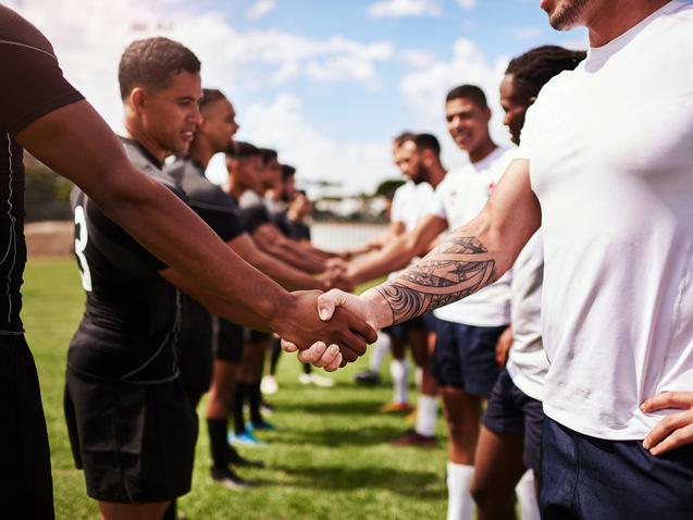 Zwei Gruppen von Rugby-Spielern sch&uuml;tteln sich die H&auml;nde auf einem gr&uuml;nen Spielfeld unter blauem Himmel.