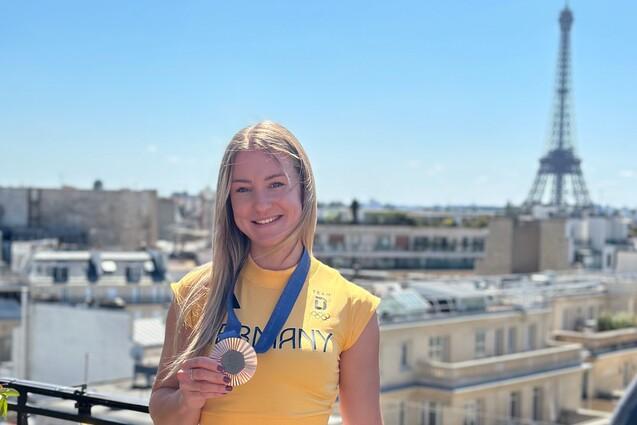 Junge Athletin in gelbem Shirt zeigt stolz ihre Medaille vor dem Eiffelturm und einem klaren blauen Himmel.