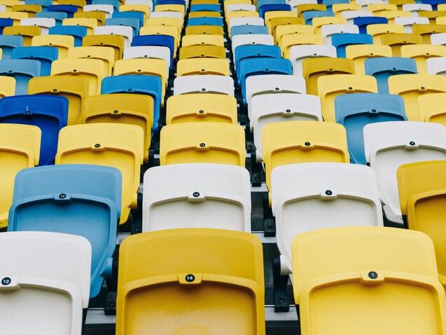 Reihe von bunten Stadionb&auml;nken in Gelb, Blau und Wei&szlig;, schr&auml;g von vorne fotografiert.