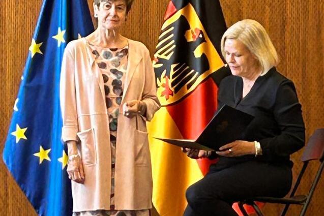 Zwei Frauen stehen auf einer B&uuml;hne, umgeben von Europaflagge und Bundesflagge, eine h&auml;lt eine Mappe.