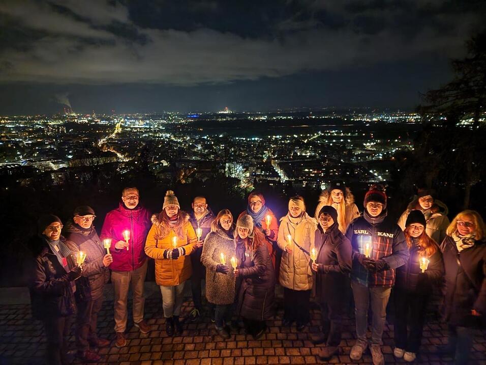 Gruppe von Menschen mit Kerzen vor einer beleuchteten Stadtlandschaft bei Nacht.