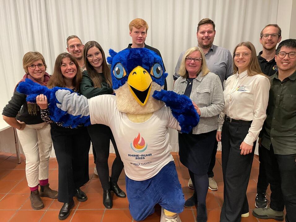Gruppe von acht Personen und einem blauen Maskottchen mit Vogelkopf, freundlich posierend vor einem wei&szlig;en Hintergrund.