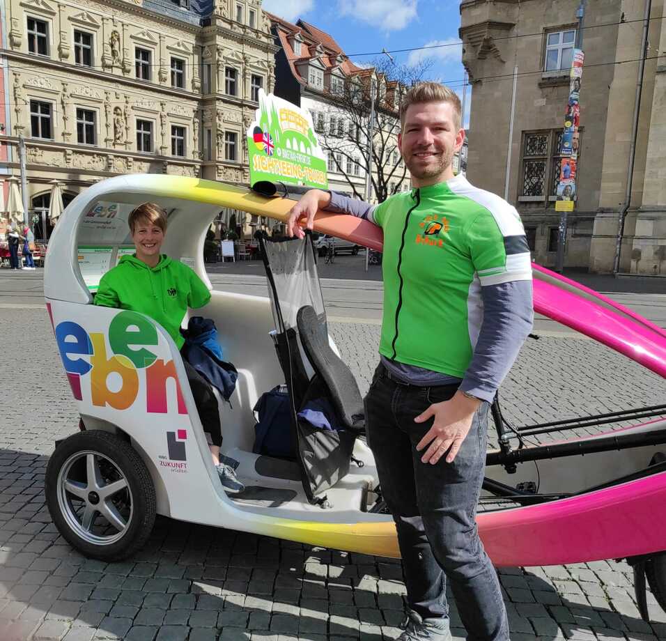 Zwei Personen in gr&uuml;nen Shirts stehen neben einem bunten Pedelac in einer Stadt, im Hintergrund historische Geb&auml;ude.