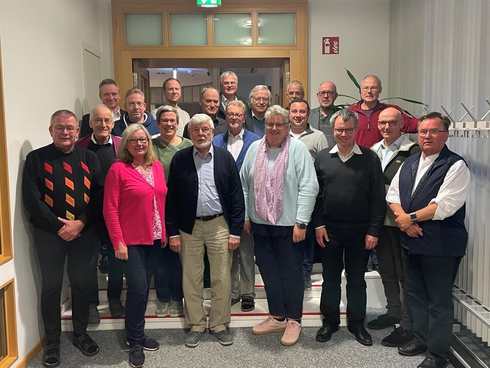 Gruppenfoto von 24 Personen in einem Flur, einige stehen, andere sitzen, alle blicken in die Kamera.