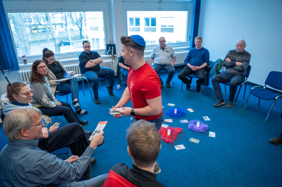 Eine Gruppe von Menschen sitzt im Kreis in einem Raum, w&auml;hrend ein junger Mann mit Kippa etwas vorzeigt.