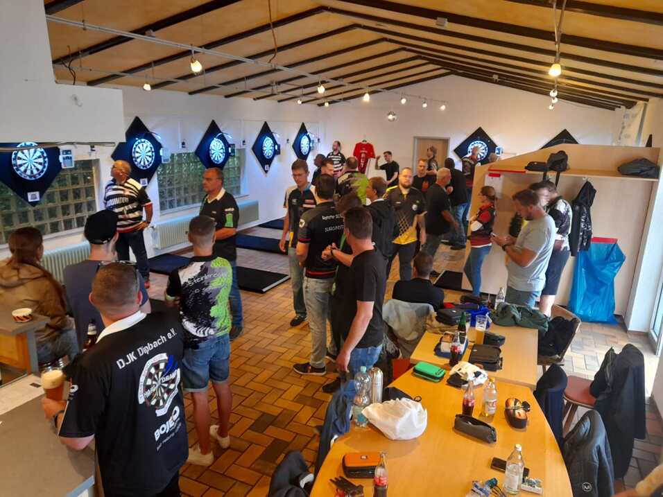 Blick in einen Dartraum mit Spielern, die sich unterhalten, Dartboards und eine bartheke im Hintergrund.