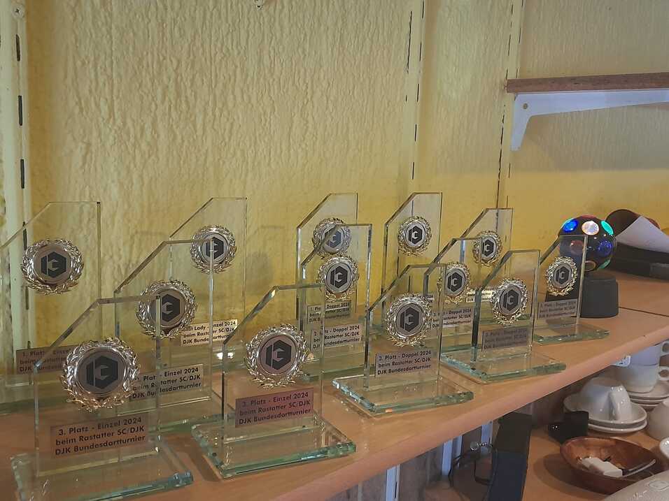 Reihe von gl&auml;sernen Pokalen mit Beschriftungen auf einem Tisch, vor einer gelben Wand.