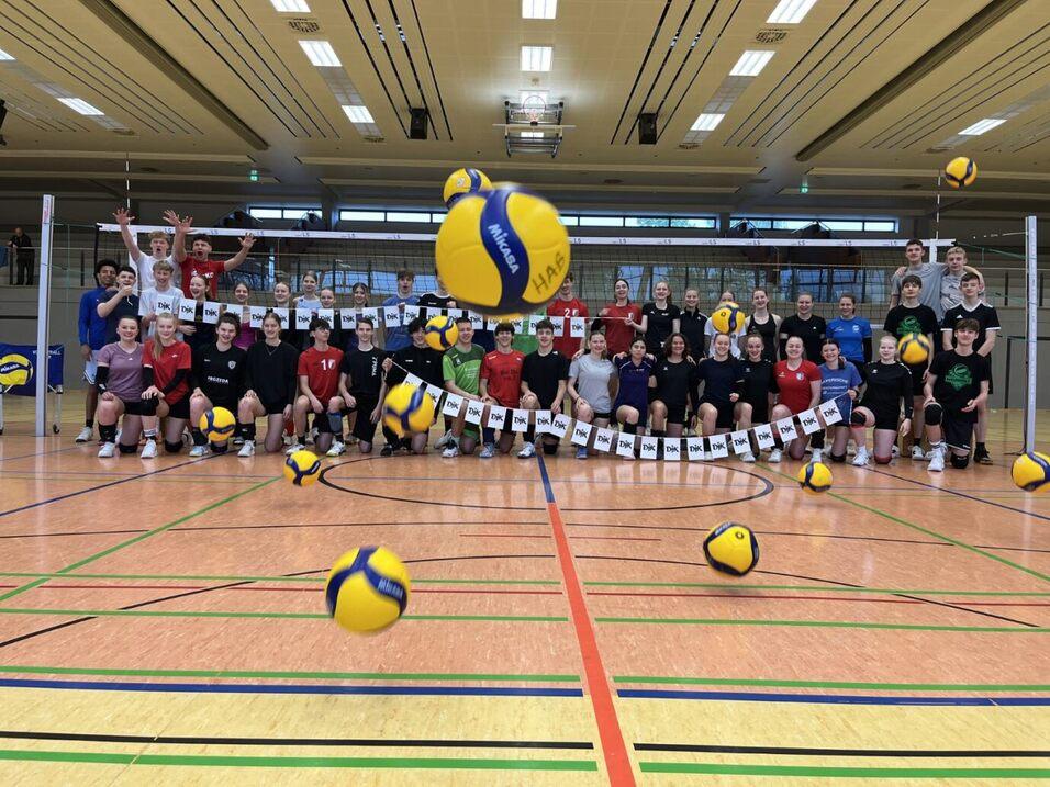 Gruppenfoto von Volleyballspielern in einer Sporthalle, mit mehreren B&auml;llen in der Luft und bunten Trikots.