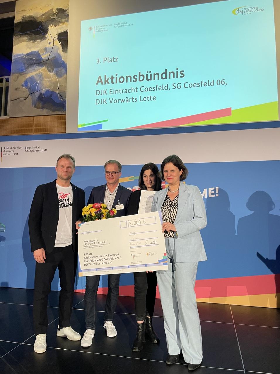 Vier Personen halten einen gro&szlig;en Scheck &uuml;ber 3.000 Euro, w&auml;hrend im Hintergrund ein Bildschirm den dritten Platz f&uuml;r &bdquo;Aktionsb&uuml;ndnis&ldquo; zeigt.