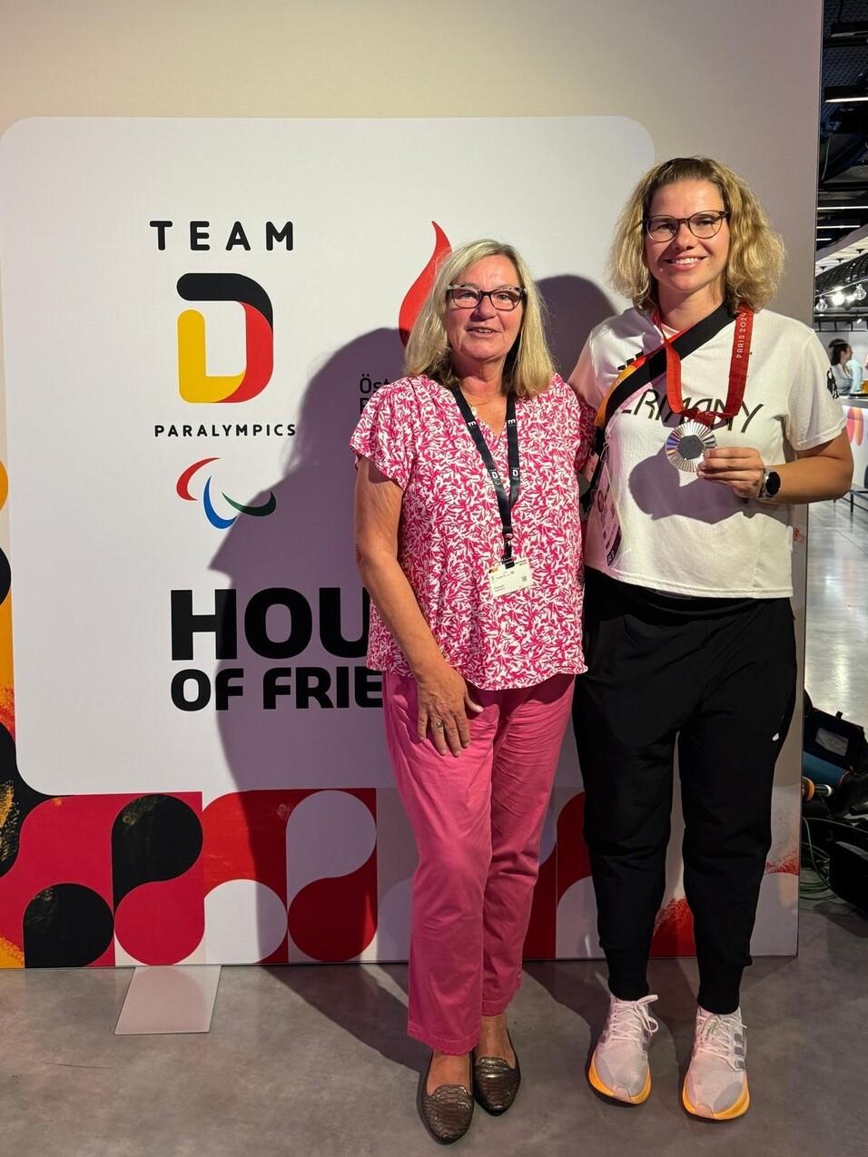 Zwei Frauen stehen vor einem bunten Hintergrund mit dem Schriftzug &bdquo;HOUSE OF FRIENDS&ldquo; und dem Paralympics-Logo.