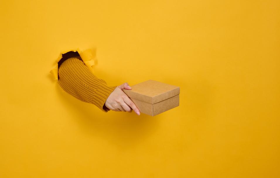 Hand in einem Loch einer gelben Wand h&auml;lt eine kleine, braune Geschenkbox.