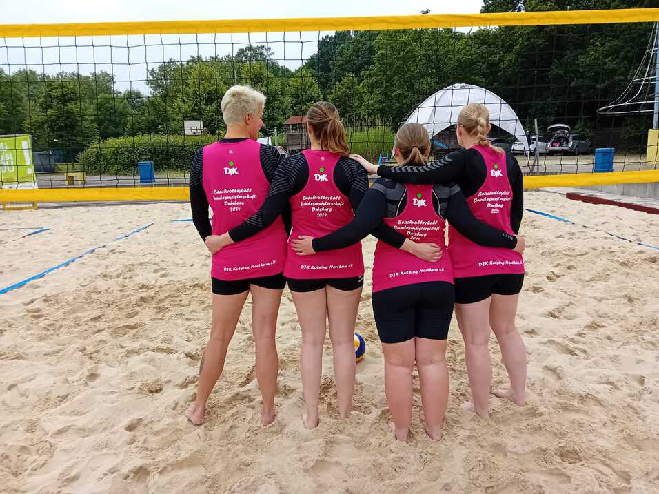 Vier Frauen in Sportkleidung stehen R&uuml;cken an R&uuml;cken am Sandstrand eines Volleyballfeldes.