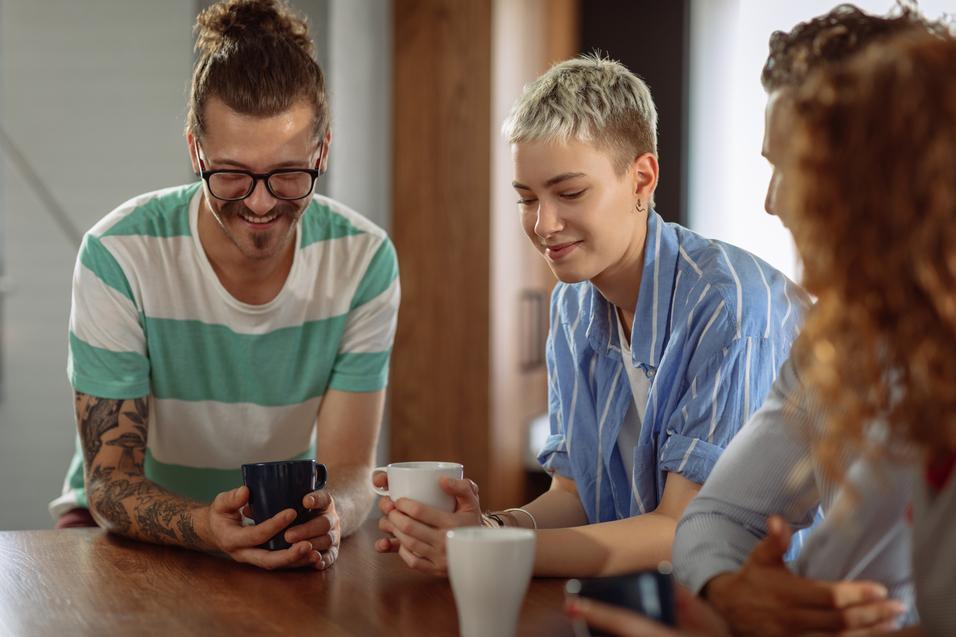Vier Personen sitzen an einem Tisch und betrachten l&auml;chelnd ein Smartphone, w&auml;hrend sie Tassen in den H&auml;nden halten.