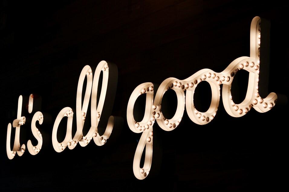 Leuchtende Schriftzug &bdquo;it's all good&ldquo; in geschwungener Schrift auf dunklem Hintergrund.