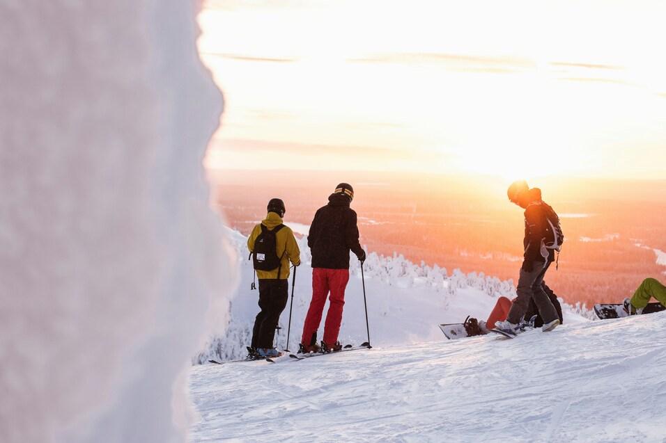Drei Skifahrer stehen auf einem schneebedeckten Hang bei Sonnenuntergang, im Hintergrund verschneite Bäume.